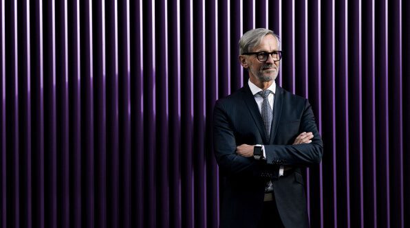 A man in a formal suit and glasses stands with arms crossed against a backdrop of purple vertical lines. He appears confident and is smiling slightly.