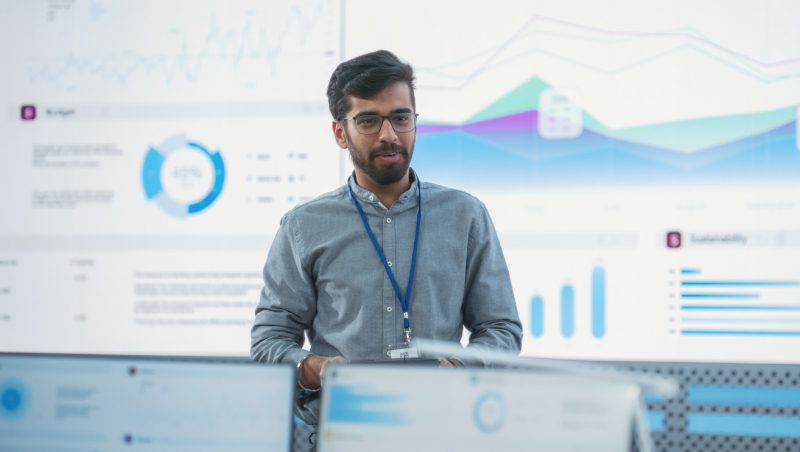 A man wearing glasses and a light blue shirt stands in front of a large screen displaying various graphs and data visuals in an office setting.