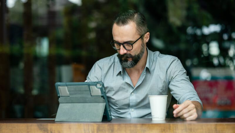 A man wearing glasses and a light gray shirt, sitting at a wooden table in a cafe, focused on a tablet. A white coffee cup is placed next to him.