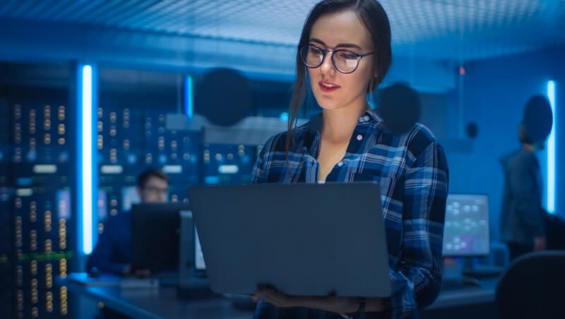 A woman in glasses and a plaid shirt working on a laptop in a server room, surrounded by blue lighting and servers. Another person is visible in the background, engaged with their computer.