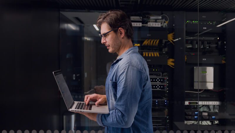 A man in a blue shirt using a laptop in a server room with rows of servers and networking equipment in the background. This setting reflects a focus on Operationalizing security and Nonstop SecOps.