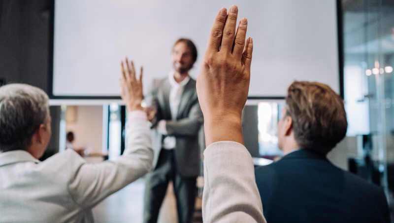 A group of three people in business attire, with two individuals raising their hands, facing a speaker in front of a screen in a modern meeting room.