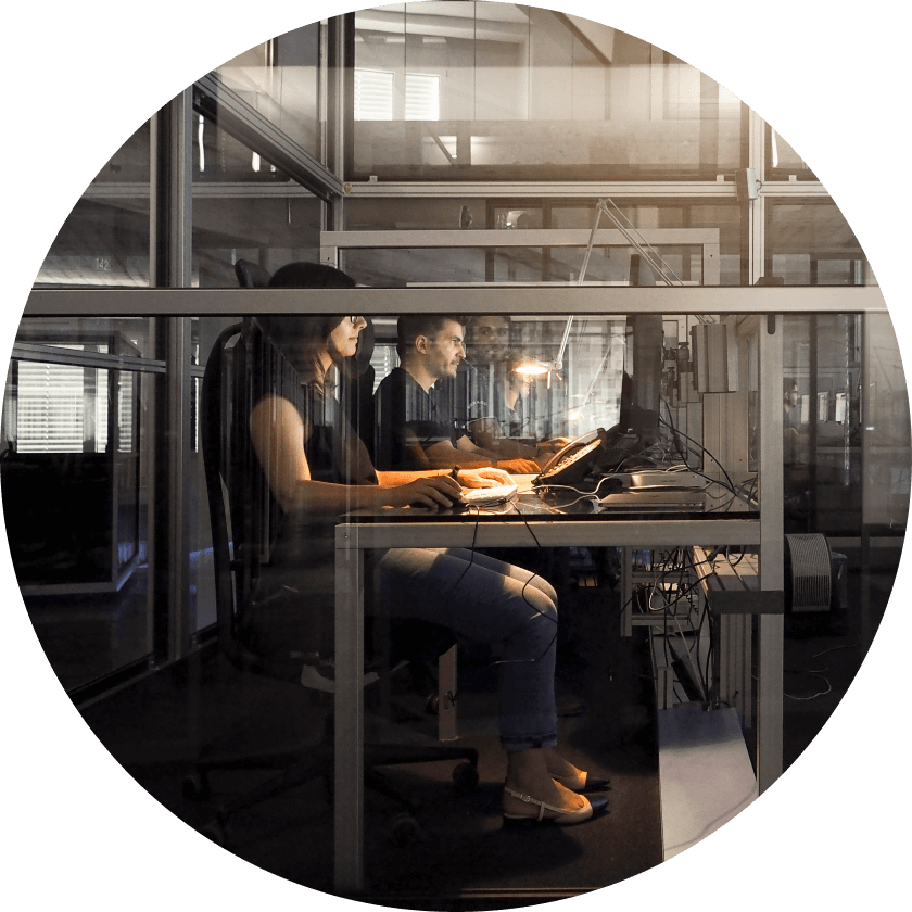 Two individuals working on computers in a modern office space with glass partitions. A woman is focused on her screen, while a man sits beside her, both illuminated by warm lighting.