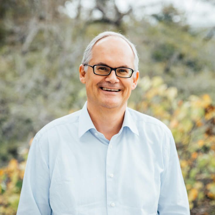 Dr. Bernd-Michael Rumpf smiling while wearing a light blue shirt, standing outdoors with blurred greenery in the background.