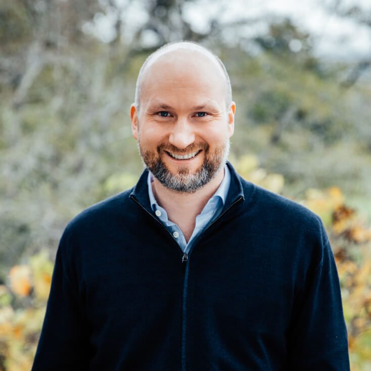 Florian Funk, wearing a dark zip-up sweater over a light blue shirt, smiles while standing outdoors with a blurred background of trees and foliage.