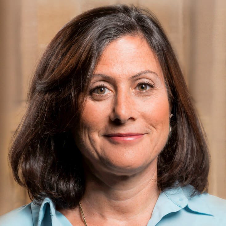 Gavriella Schuster wearing a light blue blouse, looking directly at the camera with a neutral expression. The background is soft and blurred, suggesting a professional setting.