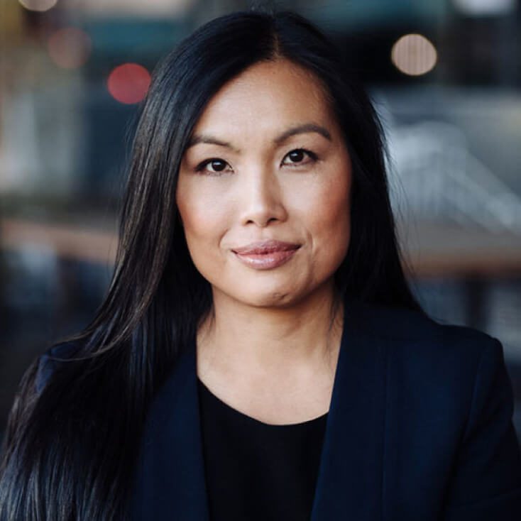 Maggie Chan Jones wearing a dark blazer, looking directly at the camera with a neutral expression. The background is blurred, featuring outdoor lighting and an urban setting.