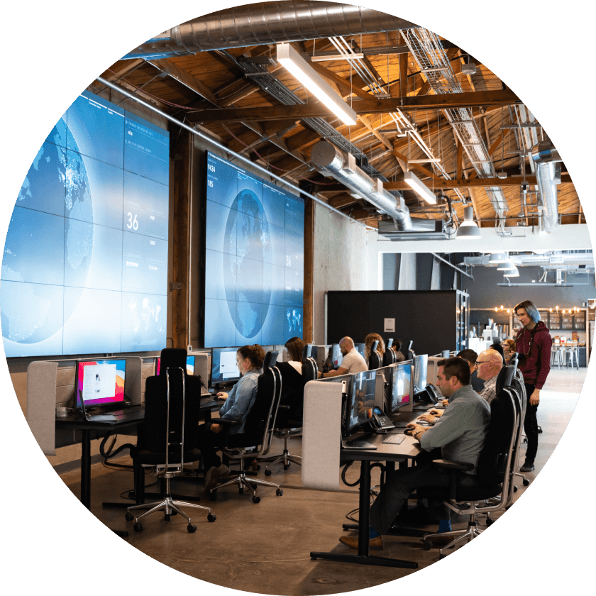 A team of professionals working at computers in a modern office, with large screens displaying data and maps in the background. The workspace features a mix of desks and chairs.