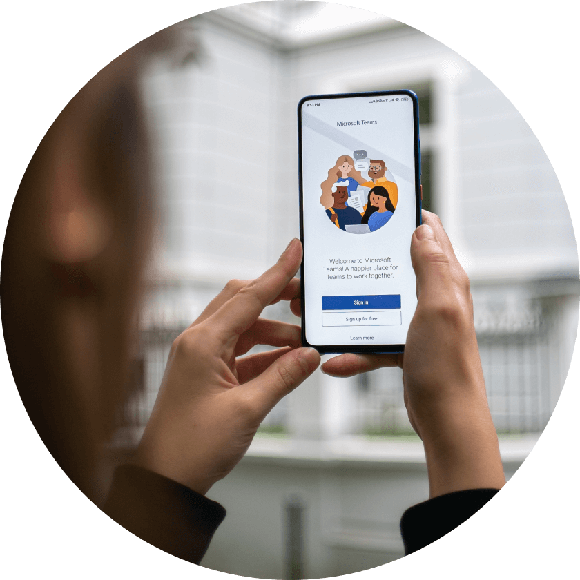 A person holding a smartphone displaying the Microsoft Teams welcome screen, featuring illustrations of diverse people collaborating. The background shows a white building.