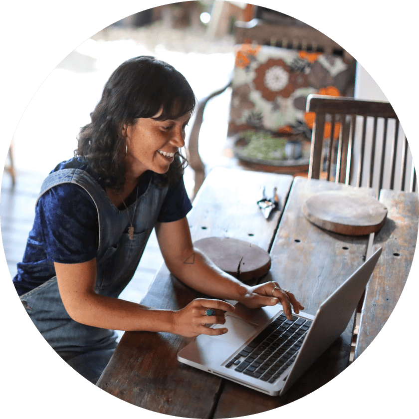 A woman sitting at a wooden table using a laptop, smiling while engaged in her work. She is wearing a blue shirt and denim overalls, with a cozy interior visible in the background.