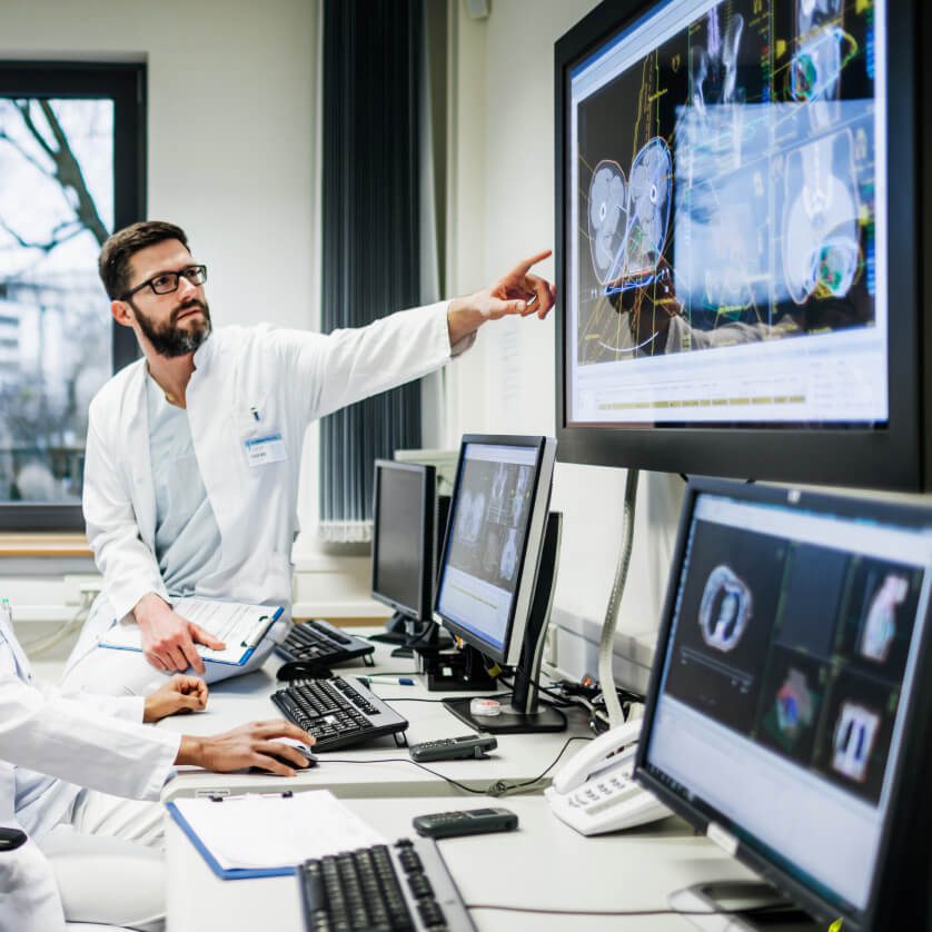 A male doctor in a white coat pointing at a medical imaging screen while speaking with a colleague seated at a computer in a modern medical office.