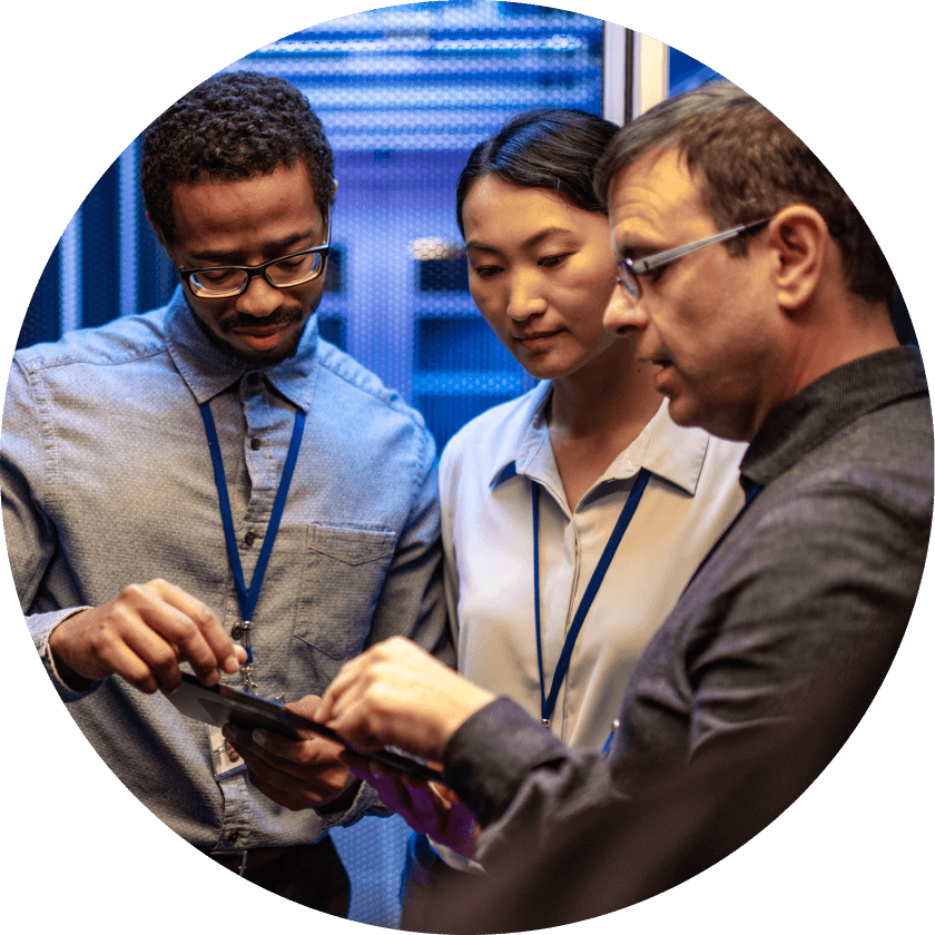 Three professionals collaborating while reviewing information on a tablet in a tech environment. Background features blue lighting and server equipment.