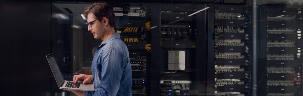 A technician working on a laptop in a server room, with network cables and servers visible in the background, emphasizing operationalizing security and nonstop SecOps.