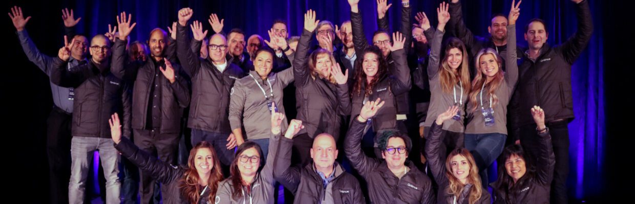 A large group of people smiling and raising their hands during an event, wearing matching jackets. The backdrop features blue lighting, creating a celebratory atmosphere. Open Systems MDR is Ontinue.