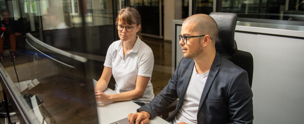 Two professionals sitting at a desk in an office environment. One person in a white shirt looks at the computer screen while the other, dressed in a dark blazer, interacts with a laptop.