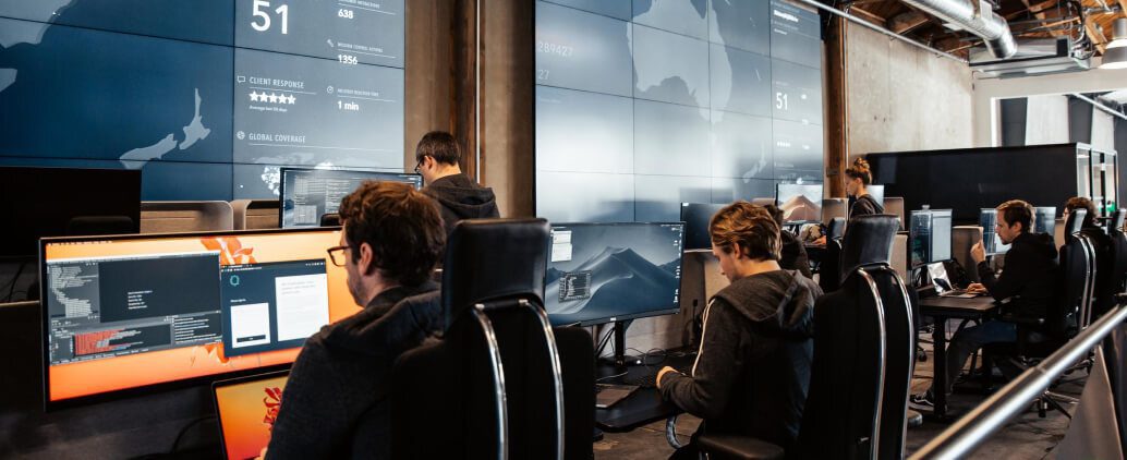 A modern office with several people working at computer desks, focused on screens displaying data and analysis, with large video walls in the background showcasing maps and statistics.