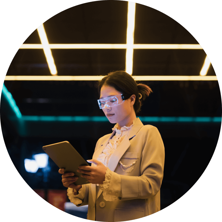 A woman wearing clear protective glasses holds a tablet while standing under bright, modern lighting. She is dressed in a light-colored blazer and is focused on the device.