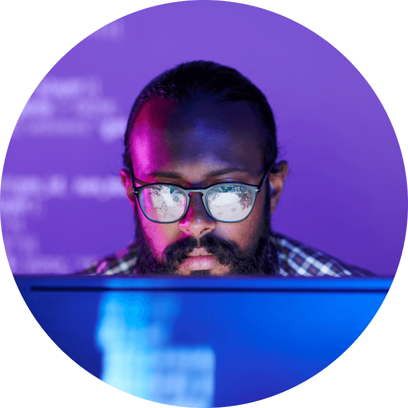 A person with glasses and a beard focused on a computer screen displaying code, with a purple background illuminated by various colors.