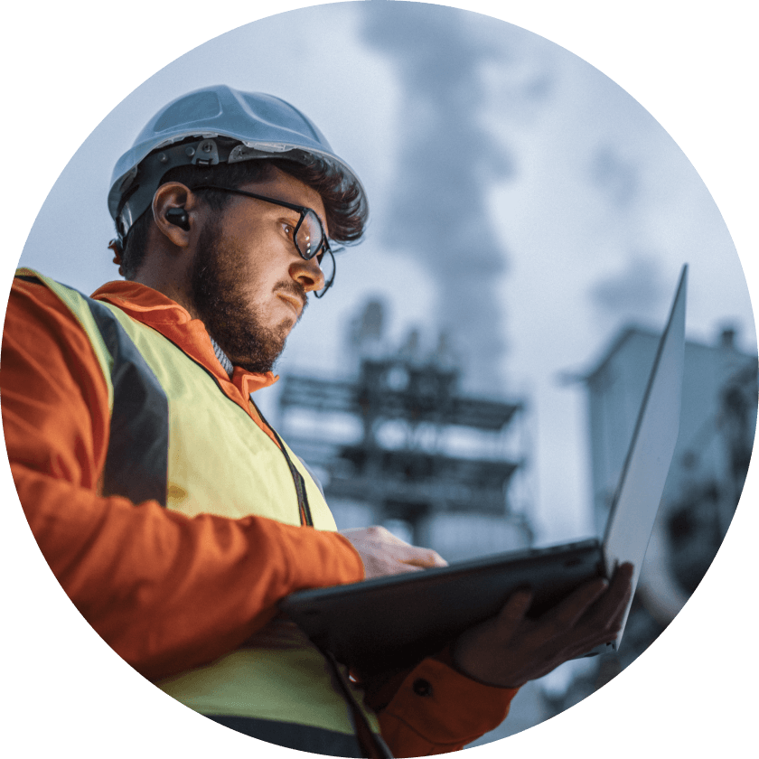 A construction worker wearing a hard hat and safety vest, using a laptop outdoors. In the background, industrial structures and smoke are visible, indicating a worksite environment.