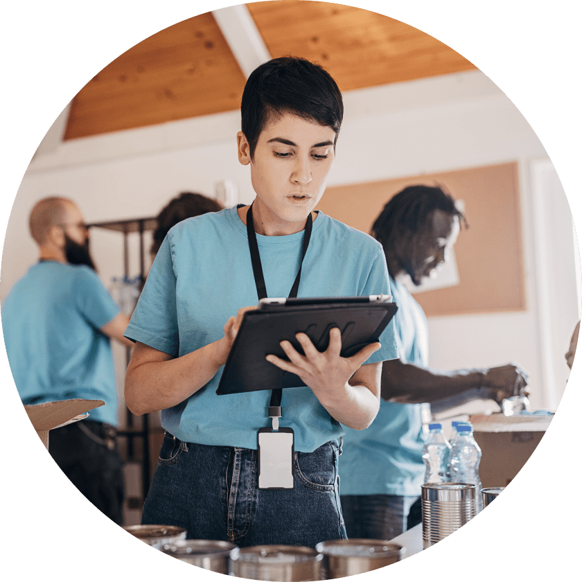 A young woman in a blue shirt holding a clipboard while concentrating on her task. In the background, other volunteers are organizing items on tables, working in a bright room.
