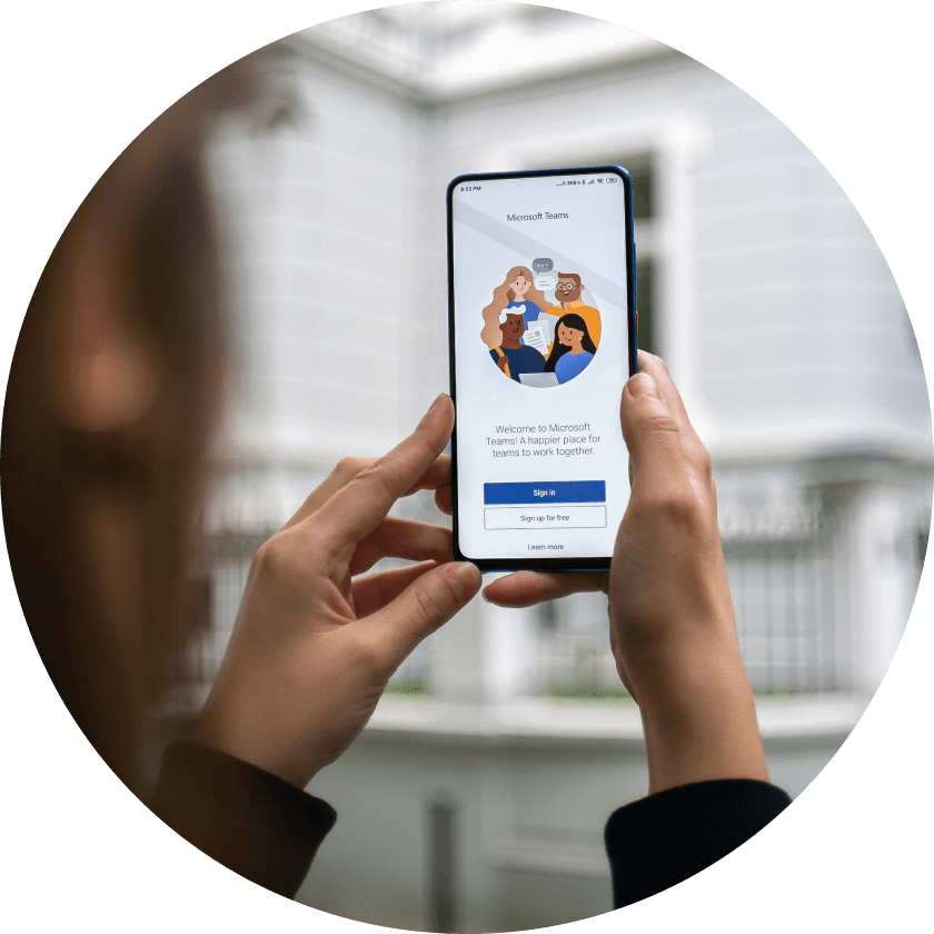 A person holding a smartphone displaying the Microsoft Teams welcome screen, featuring illustrations of diverse team members and options to sign in or sign up.