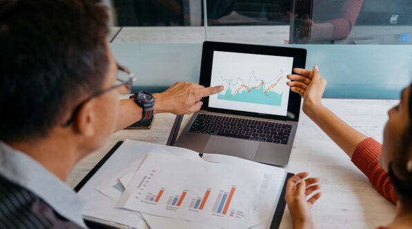 Two professionals discussing data during a meeting. One person is pointing at a laptop screen displaying graphs, while the other is gesturing toward the screen with printed reports on the table.