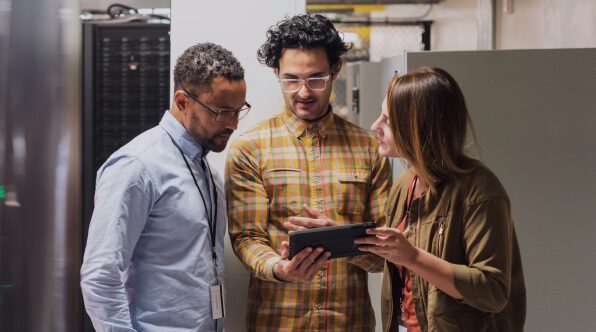 Three colleagues discussing a project while looking at a tablet in a modern workspace. The individuals include two men and one woman, all dressed in casual attire.