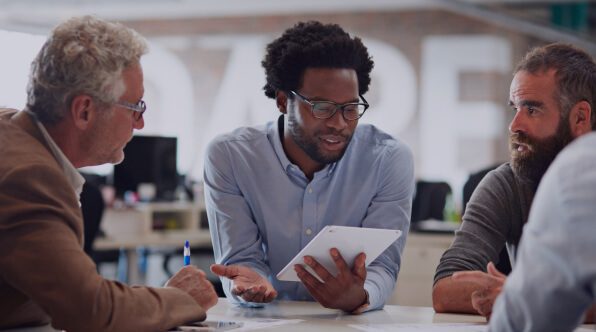 A diverse group of four professionals engaged in a discussion around a table, with one man holding a tablet, while the others listen attentively in a modern office environment. The setting emphasizes collaboration and teamwork.