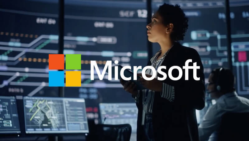 A woman standing in a data analytics room, looking at digital screens displaying technical graphs and information, with a person sitting at a desk in the background. The Microsoft logo is displayed prominently.
