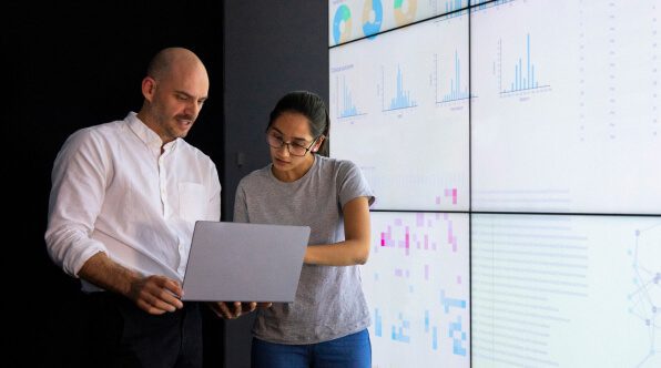 A man and a woman discussing data on a laptop in front of digital screens displaying various graphs and charts. The environment appears modern and analytical, focusing on data analysis and teamwork to reduce Microsoft Sentinel data costs.