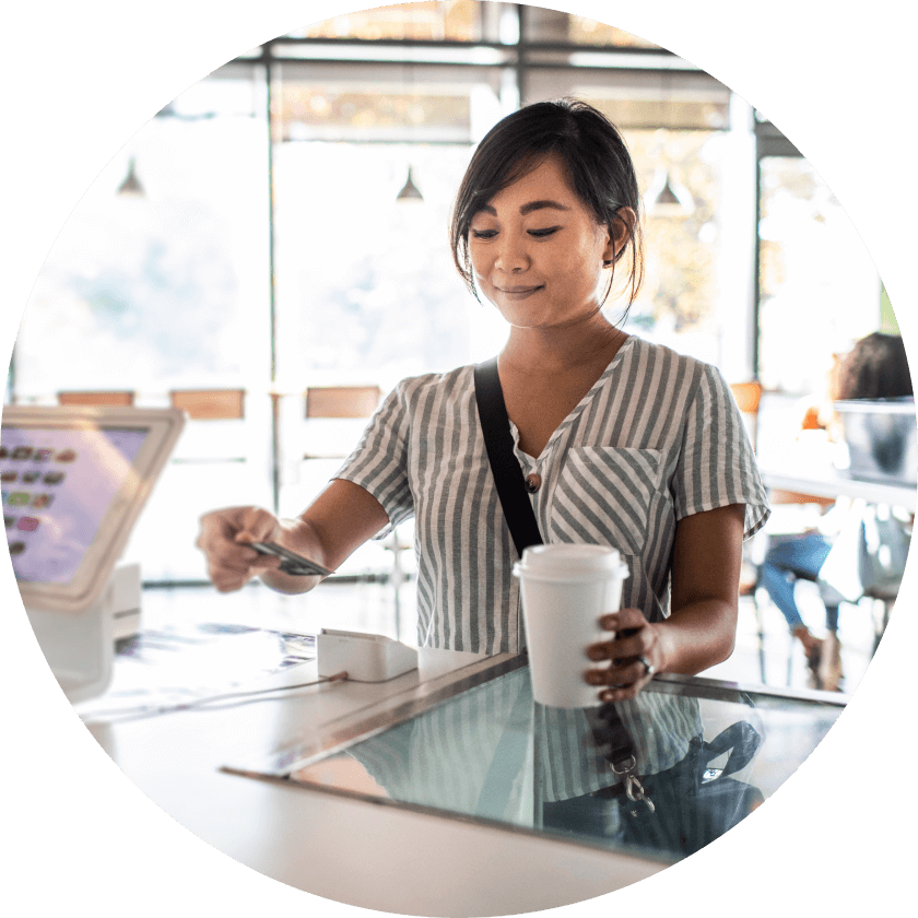 A woman in a striped shirt standing at a cafe counter, using a card to pay while holding a coffee cup. The interior features large windows and soft lighting.