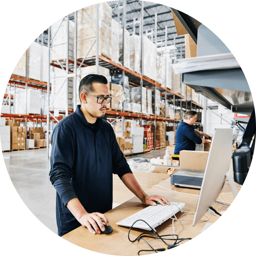 A man with glasses working at a computer in a warehouse with shelves stocked with boxes in the background. Another worker is visible in the background, organizing items.