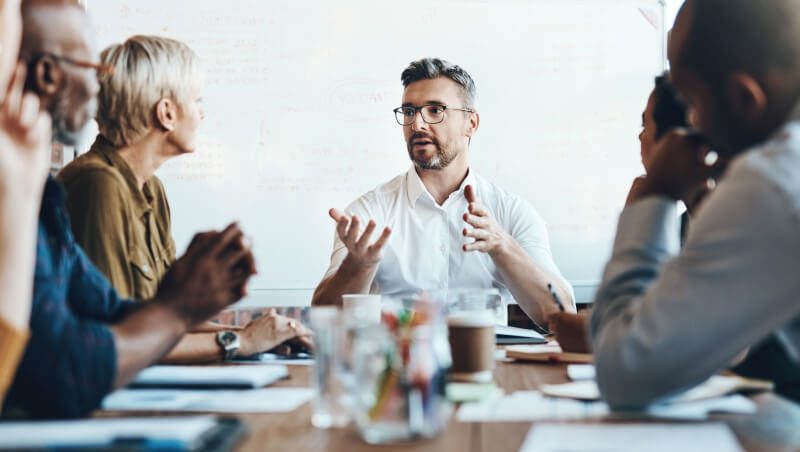 A group of professionals engaged in a meeting around a wooden table, with a man speaking and gesturing, while others listen attentively, with papers and cups visible on the table.