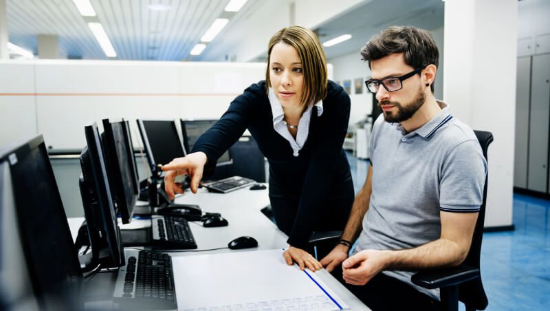 A woman pointing at a computer screen while explaining something to a man seated next to her in an office environment. Multiple computers are visible in the background.