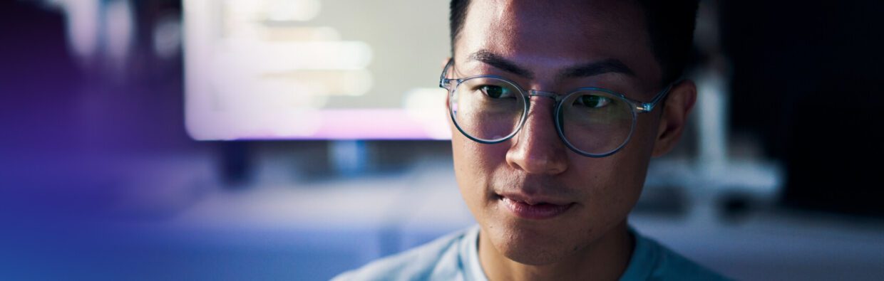 A person with glasses focused on a screen displaying code, seated in a dimly lit room. The background is slightly blurred, emphasizing the individual’s concentration and work environment.