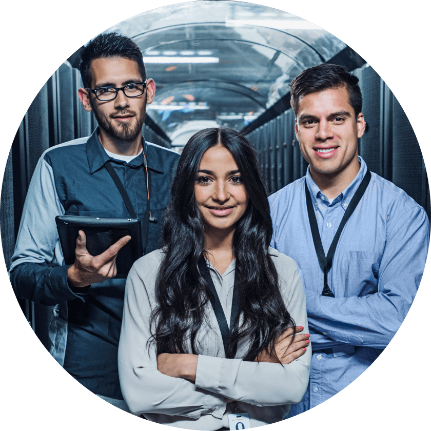Three professionals posing confidently in a data center. The woman in the center has long hair and is wearing a light-colored blouse, flanked by two men, one holding a tablet and the other in a button-up shirt.