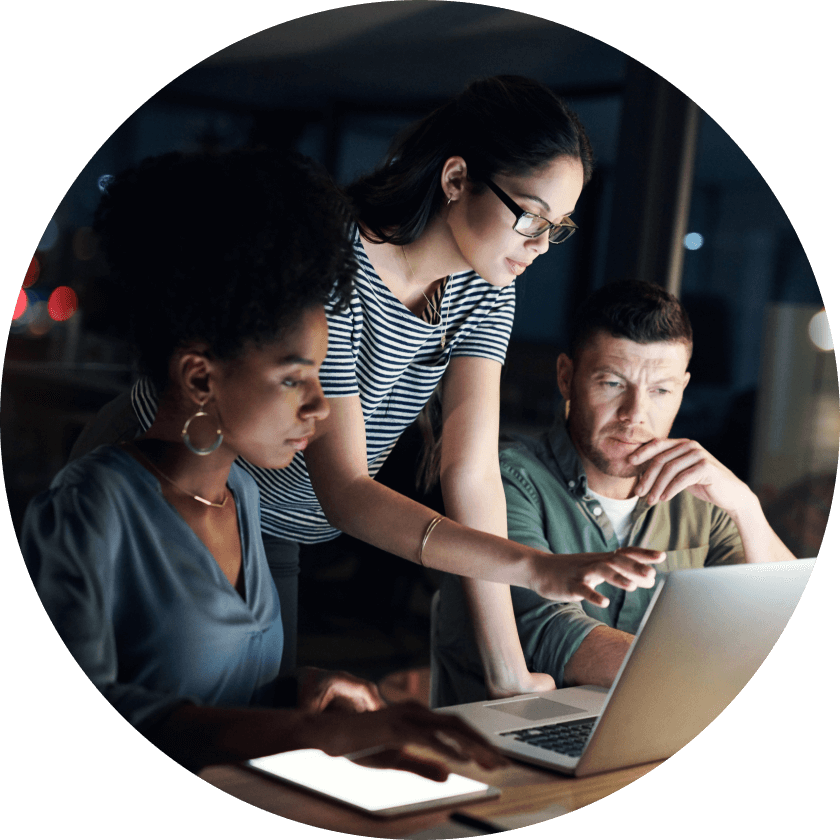 A group of three colleagues collaborating on a laptop at a desk in a dimly lit office. One woman is pointing at the screen while another woman and a man observe.