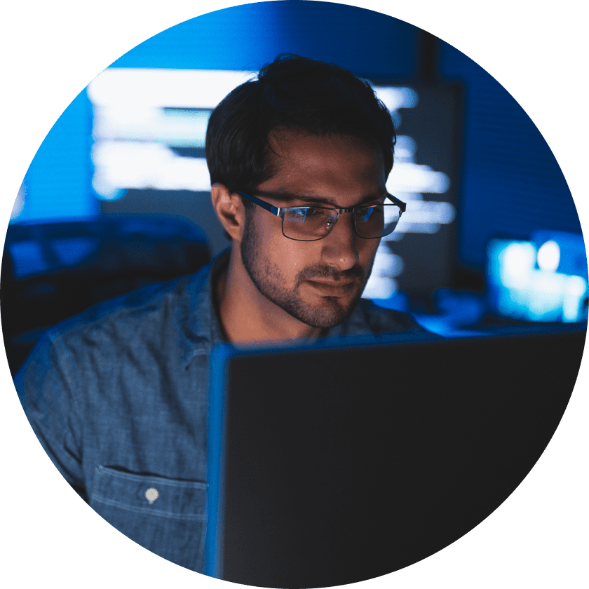A man with glasses focused on a computer screen, working in a dimly lit room with blue lighting and digital code in the background.