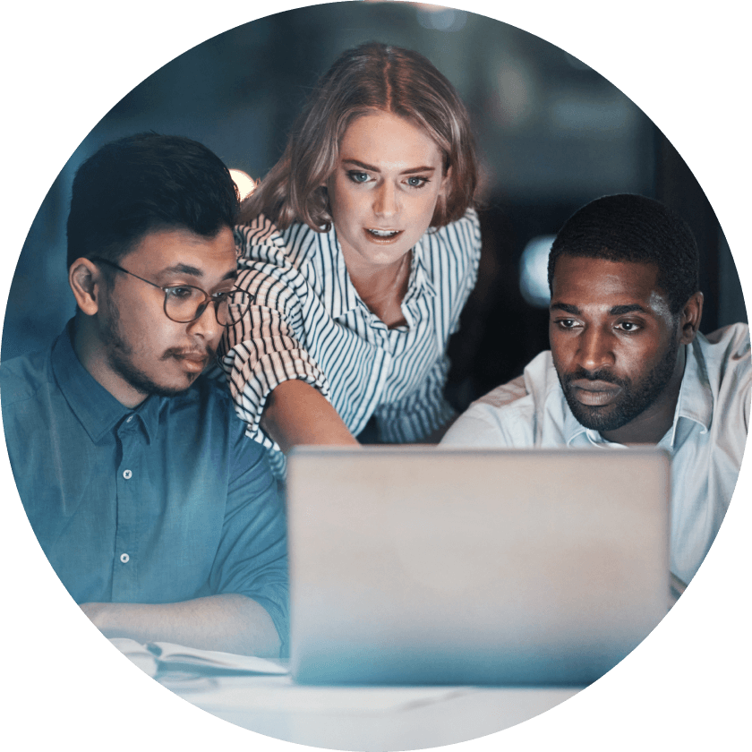 Three colleagues engaged in a discussion while looking at a laptop screen in a dimly lit environment.