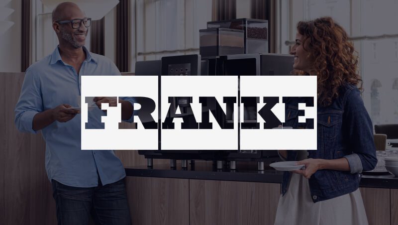 A man and woman engaging in conversation near a coffee machine in a modern kitchen setting. The logo 'FRANKE' is prominently displayed across the image.