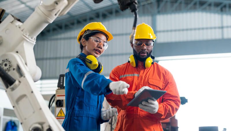 Two workers discussing technical details in a factory setting. One is wearing a blue work uniform and yellow helmet, while the other is in an orange jumpsuit and yellow helmet. A robotic arm is visible in the background.