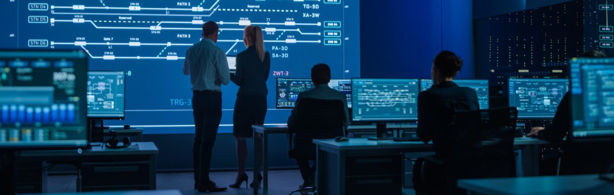 Two professionals discussing information in front of a large digital display, surrounded by several monitors showing data analysis in a modern control room.