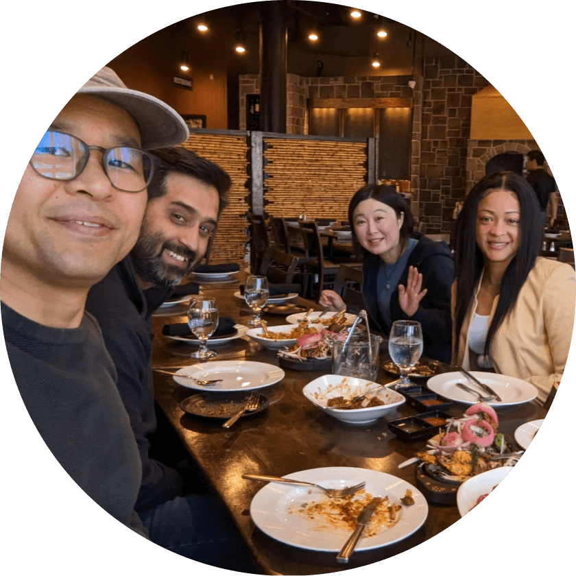 A group of four people sitting at a dining table in a restaurant, smiling and posing for the camera. Plates with leftover food and drinks are visible on the table.
