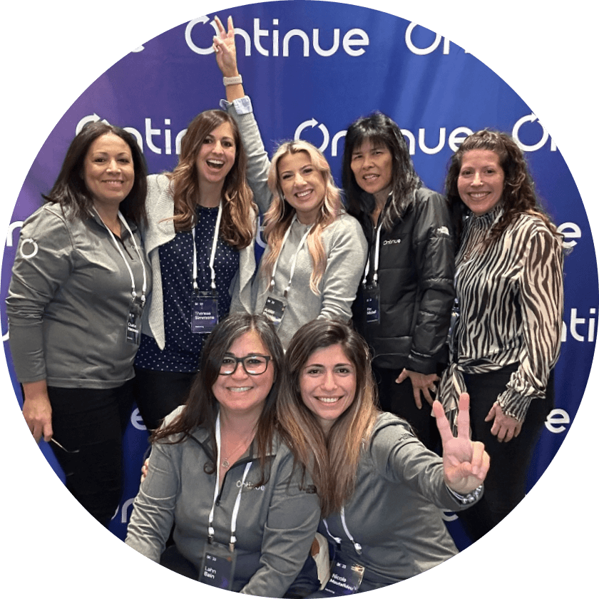 A group of seven women posing for a photo in front of a blue backdrop with the word 'Continue' in white. The women are wearing matching gray jackets and are smiling, with one woman giving a peace sign.