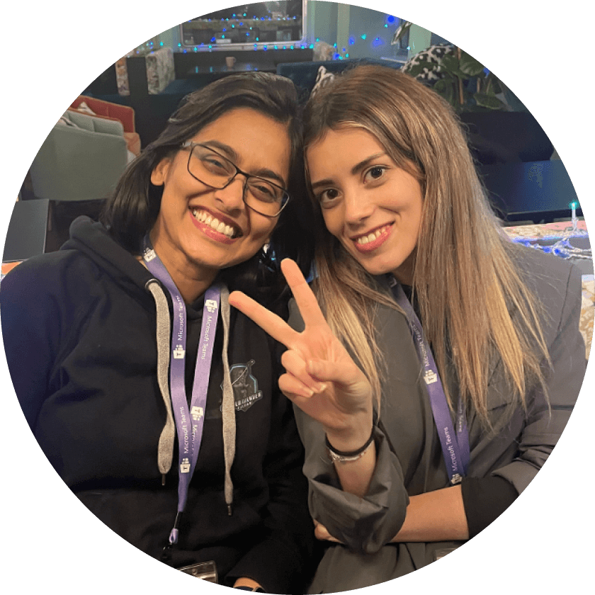 Two women smiling while sitting close together, one making a peace sign with her hand. Both are wearing lanyards and casual attire, with a festive background featuring soft lighting.
