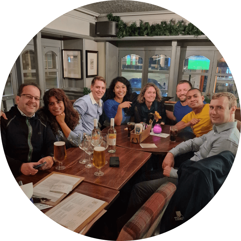 A group of eight people gathered around a wooden table in a pub, enjoying drinks and food. The setting includes glasses of beer, bottles, and a menu. The atmosphere appears relaxed and social.