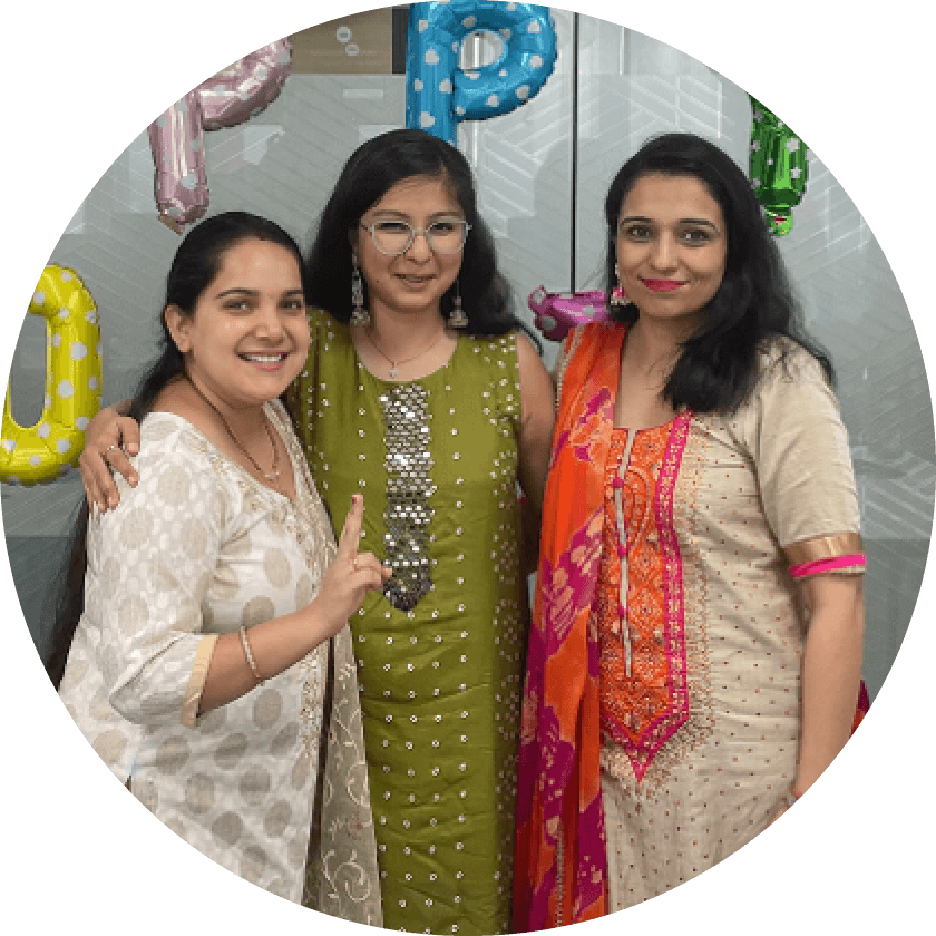 Three women posing together at a celebration, wearing traditional attire. They smile and stand in front of colorful balloon decorations spelling out 'UP'. The background features a modern office interior.