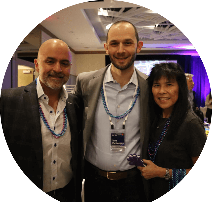 Three people posing together at an event, smiling. One man on the left has short, dark hair and is wearing a gray jacket, while the man in the middle has a beard and is dressed in a light shirt. A woman on the right has long black hair and is wearing blue accessories.