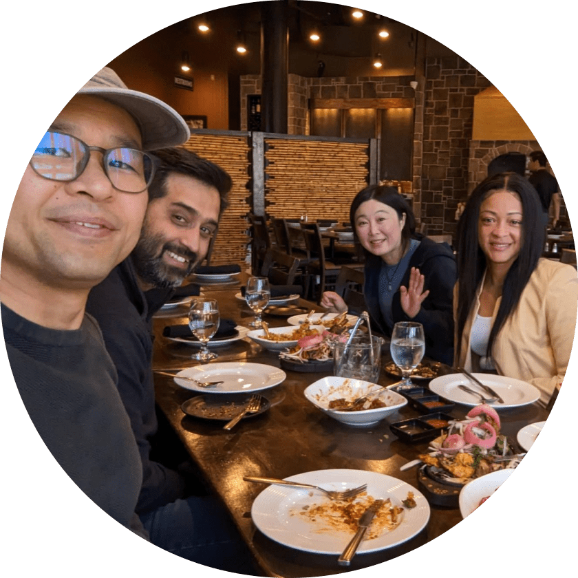 Four people seated at a table in a restaurant, smiling and posing for the camera. The table is set with empty plates and glasses, indicating they have finished their meal. The restaurant has a warm ambiance with wooden decor.