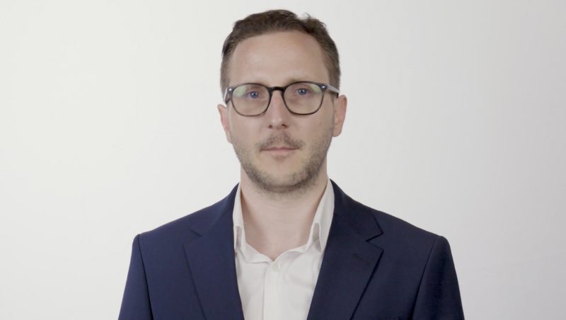 A man wearing glasses and a dark suit stands against a plain white background. He has short hair and a neutral expression.